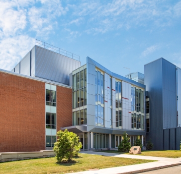 A multi-faceted building with a mix of brick, metal, and glass panels stands under a bright sky with scattered clouds, showcasing a blend of architectural styles.