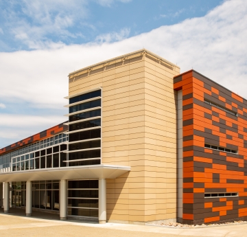 A contemporary building with a geometric design, featuring a tall beige section with rectangular windows and a contrasting section with an orange and brown pixelated pattern, under a partly cloudy sky.