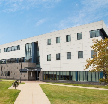 The Technology Building at Buffalo State University, a modern structure with white panels and dark windows, stands under a bright sky with a paved walkway leading to its entrance.