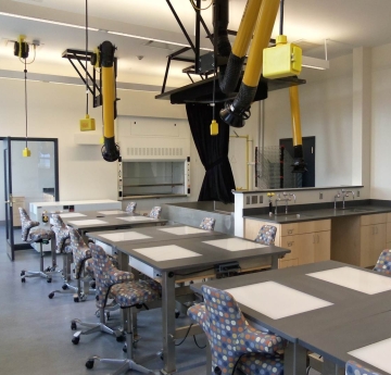 A bright, empty classroom features rows of tables with patterned chairs, overhead yellow art conservation equipment, and a sink area along the back wall