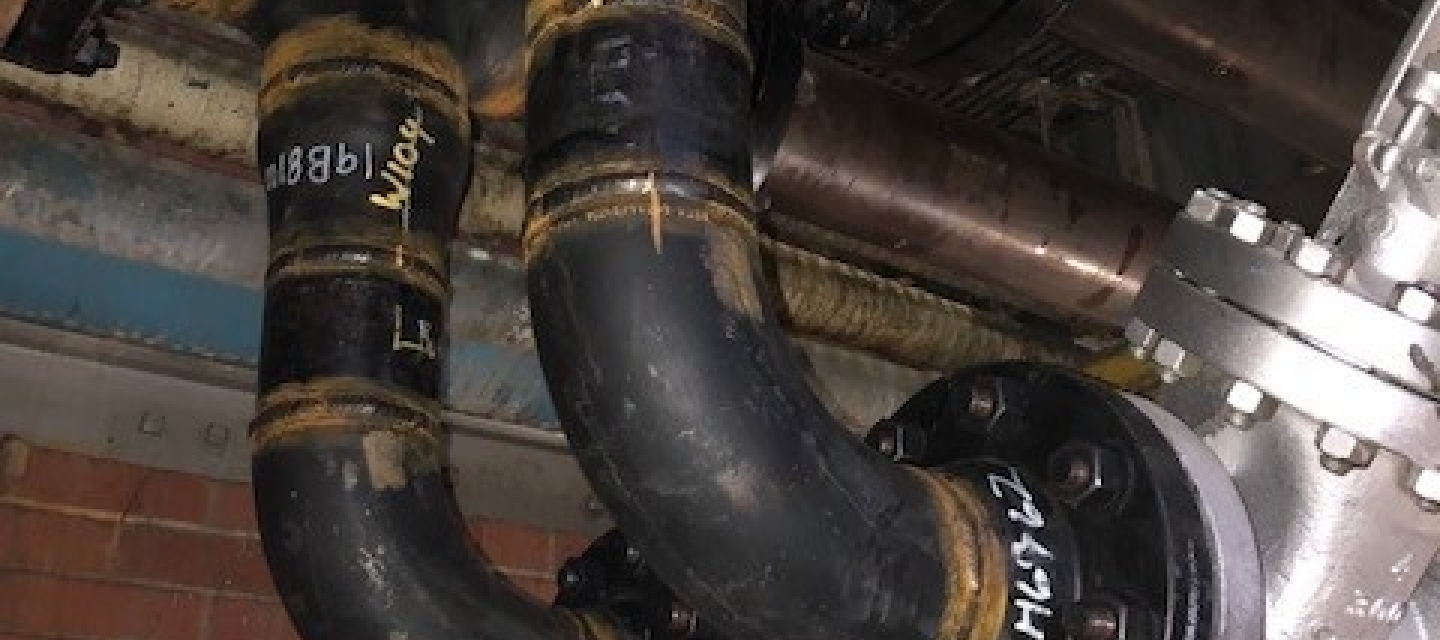 Two large, dark pipes, possibly cast iron, with rust-colored bands and various bolted connections, are shown against a backdrop of brick and other industrial piping.
