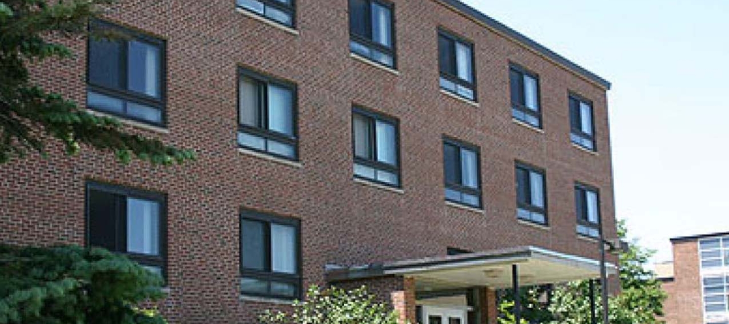  Neumann Hall, a four-story brick building with multiple windows, stands among green trees and bushes under a clear sky.